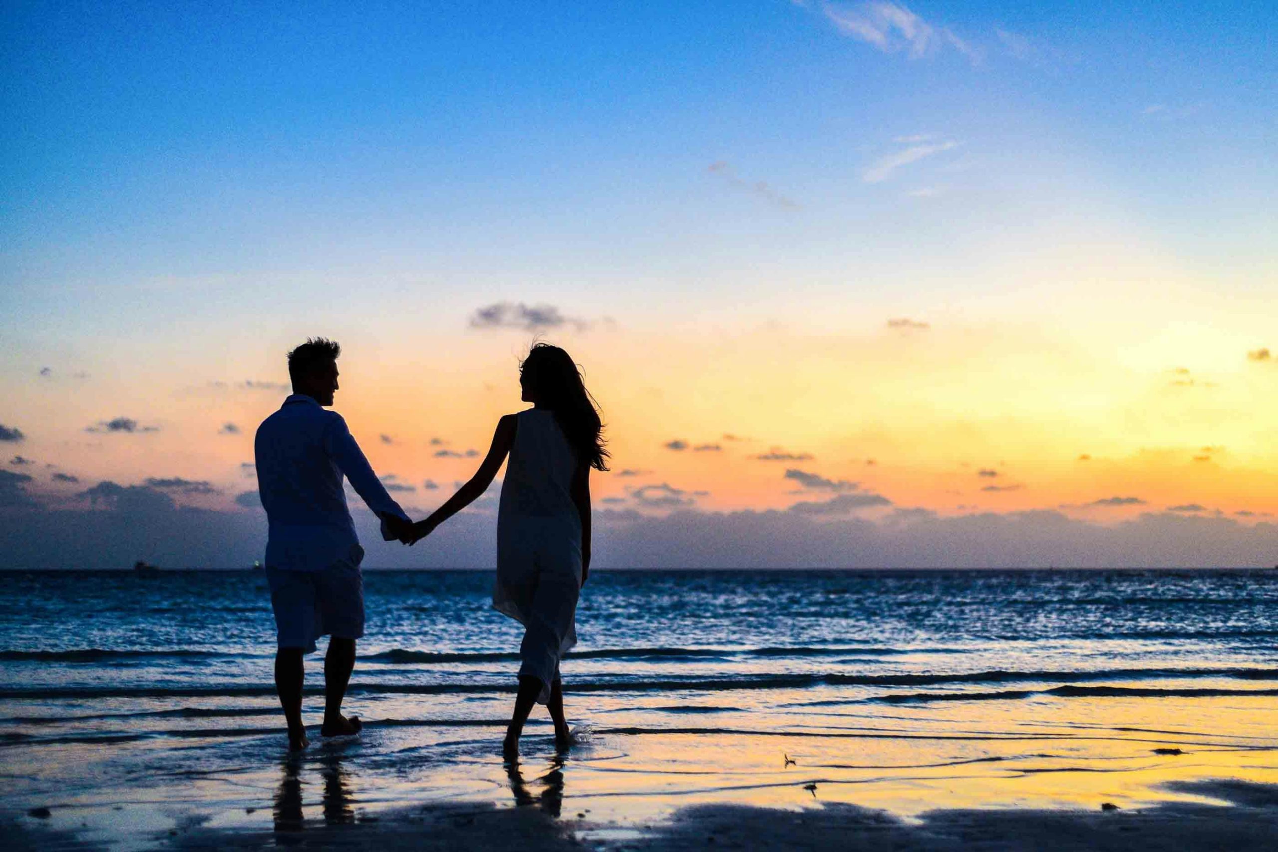 Home A romantic couple walks hand in hand on a tropical beach at sunset, enjoying a serene moment together.